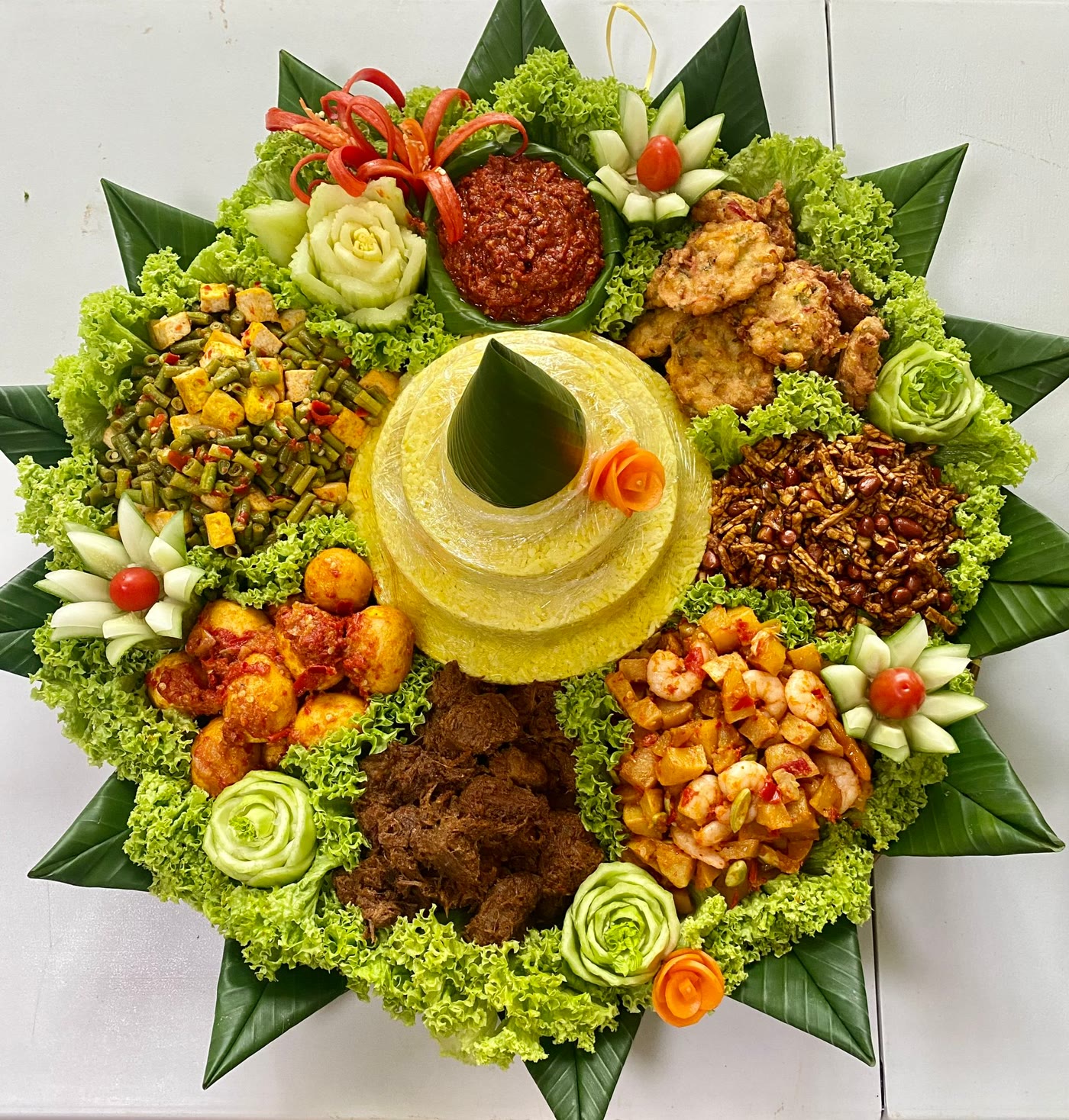 Tumpeng on a round tampah tray with yellow rice, rendang, egg balado, potato with prawn, corn fritters, tempe and sambal